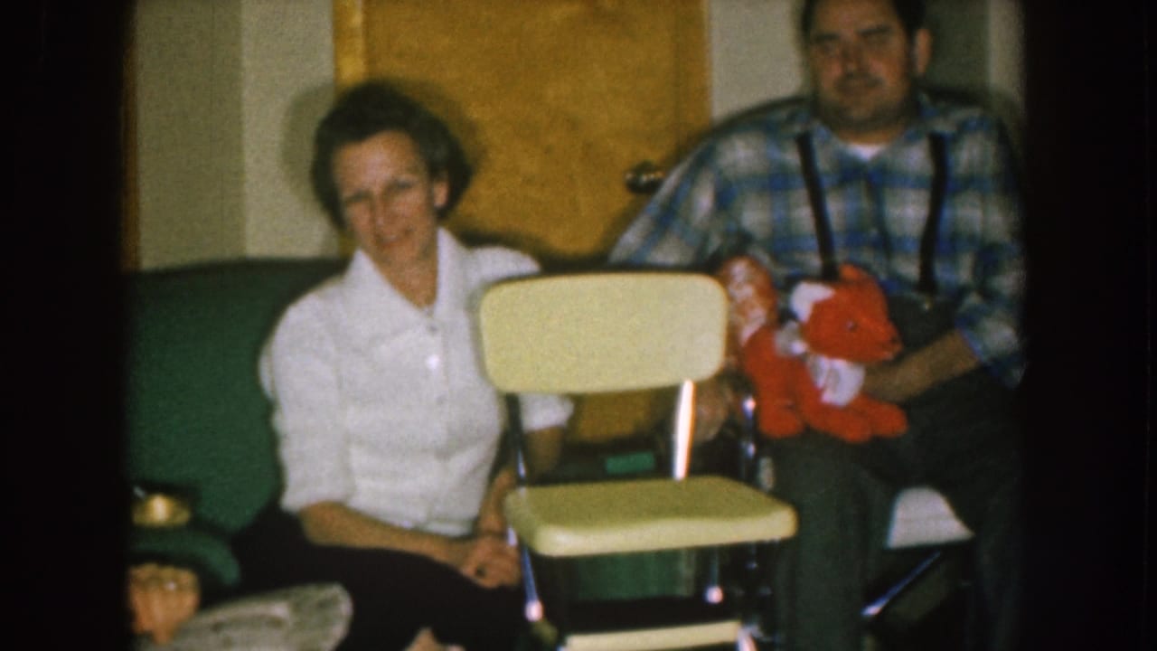Woman and man seated indoors beside a pale chair while the man holds a red stuffed animal.