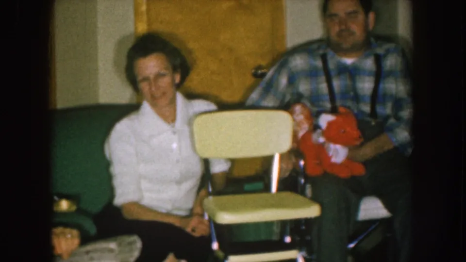 In a 1957 Nebraska home-movie frame, a woman in a white blouse sits beside a man in plaid suspenders holding a red stuffed animal.
