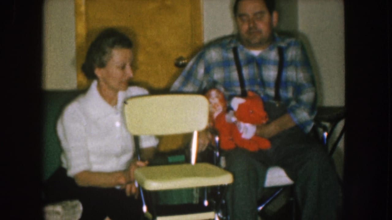 Seated woman turns toward a man holding a red stuffed animal near a pale chair.