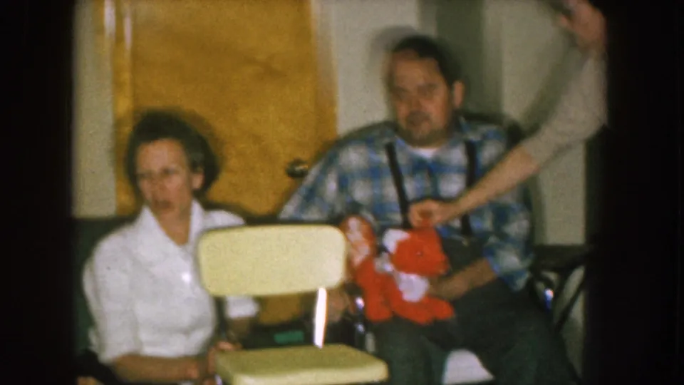 A second woman’s arm enters the frame to touch the red stuffed animal held by the seated man in the 1957 Nebraska interior.