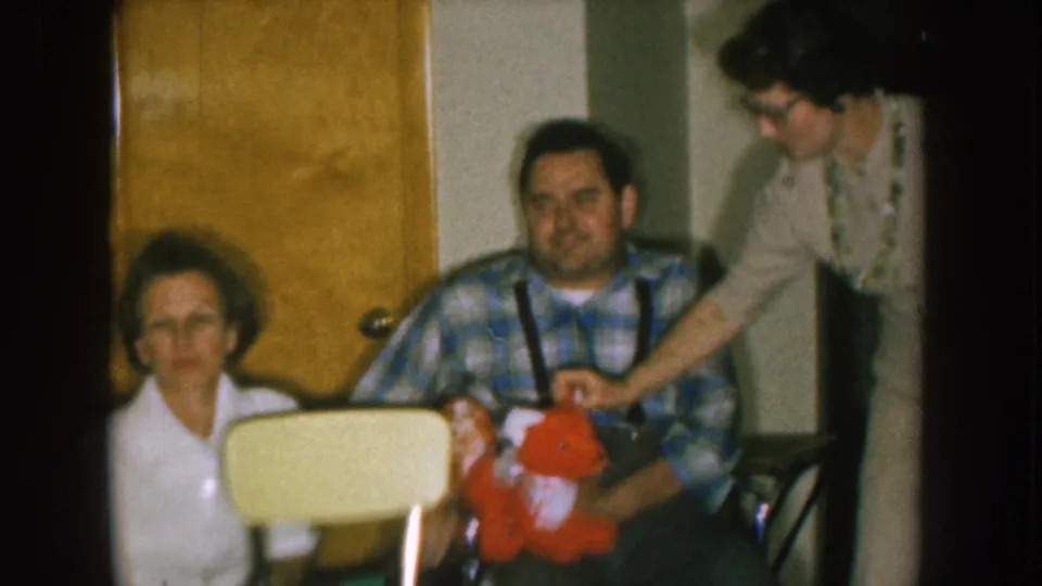 A standing woman leans over the seated pair and appears to squeeze the red stuffed animal in a 1957 Nebraska home-movie scene.