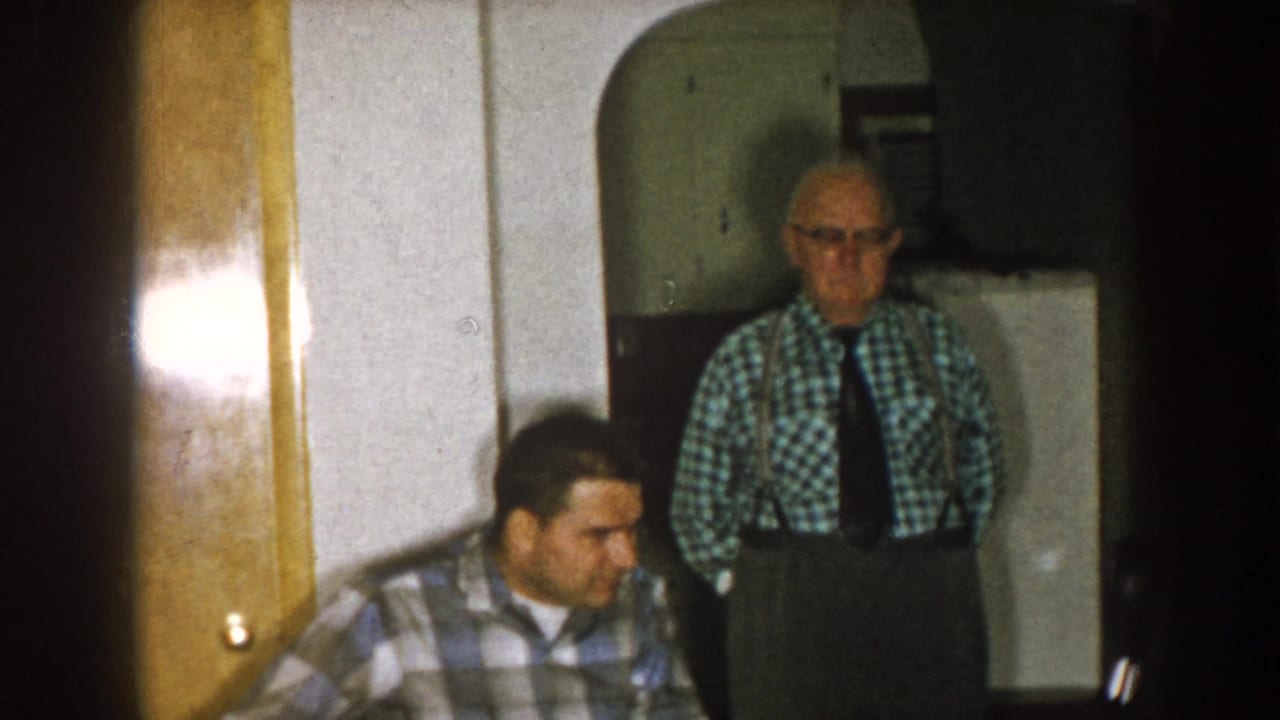 Nebraska 1957 home-movie frame showing two adult men in a dim living room, with wood doors and a standing man in suspenders.