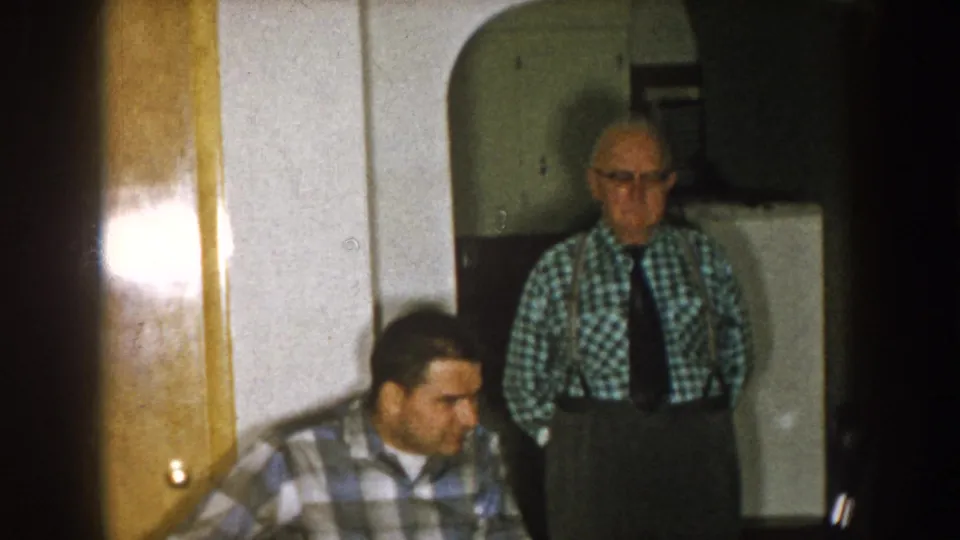Nebraska 1957 home-movie frame showing two adult men in a dim living room, with wood doors and a standing man in suspenders.