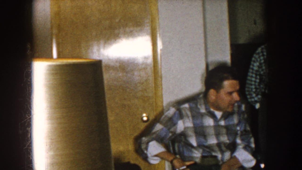A seated man in plaid appears beside a large lampshade and wood door.
