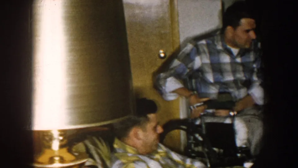 Nebraska 1957 home-movie frame showing a man seated in a wheelchair, another man in the foreground, and a shaded lamp indoors.