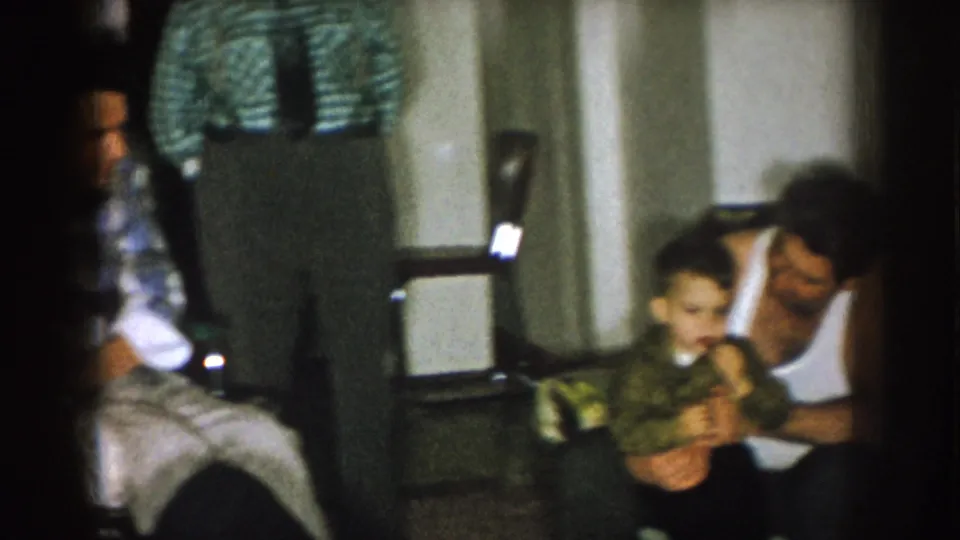 Nebraska 1957 living-room frame with adults gathered indoors and one man holding a small child near the right edge.