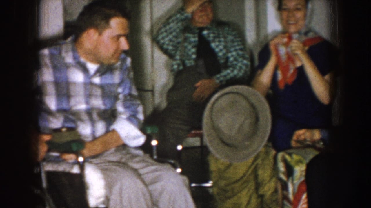 1957 Nebraska home-movie frame showing adults in a private room, including a man in a wheelchair, seated together during an informal gathering.