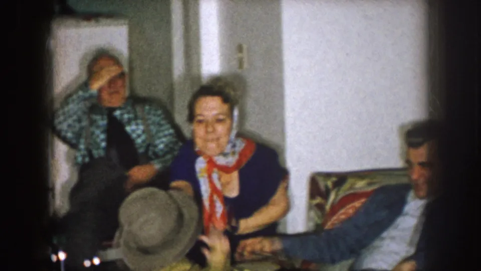 1957 Nebraska home-movie frame of adults gathered indoors, including a woman with a red scarf and men seated around her.