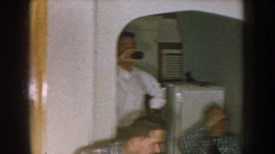 1957 Nebraska home-movie frame showing a soft, handheld view of adults near a doorway and refrigerator inside a private home.