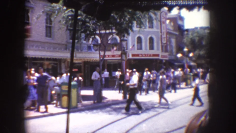 Visitors walk across a Disneyland street with streetcar tracks, storefronts, and a theater-like facade in view.