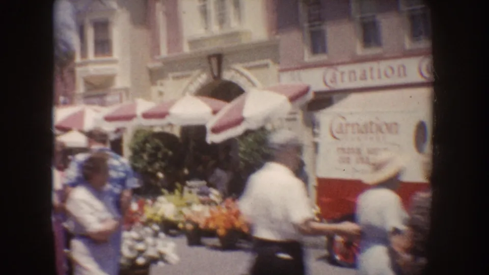 Disneyland visitors stand near striped umbrellas, flower displays, and a storefront with visible signage in a 1962 home-movie frame.