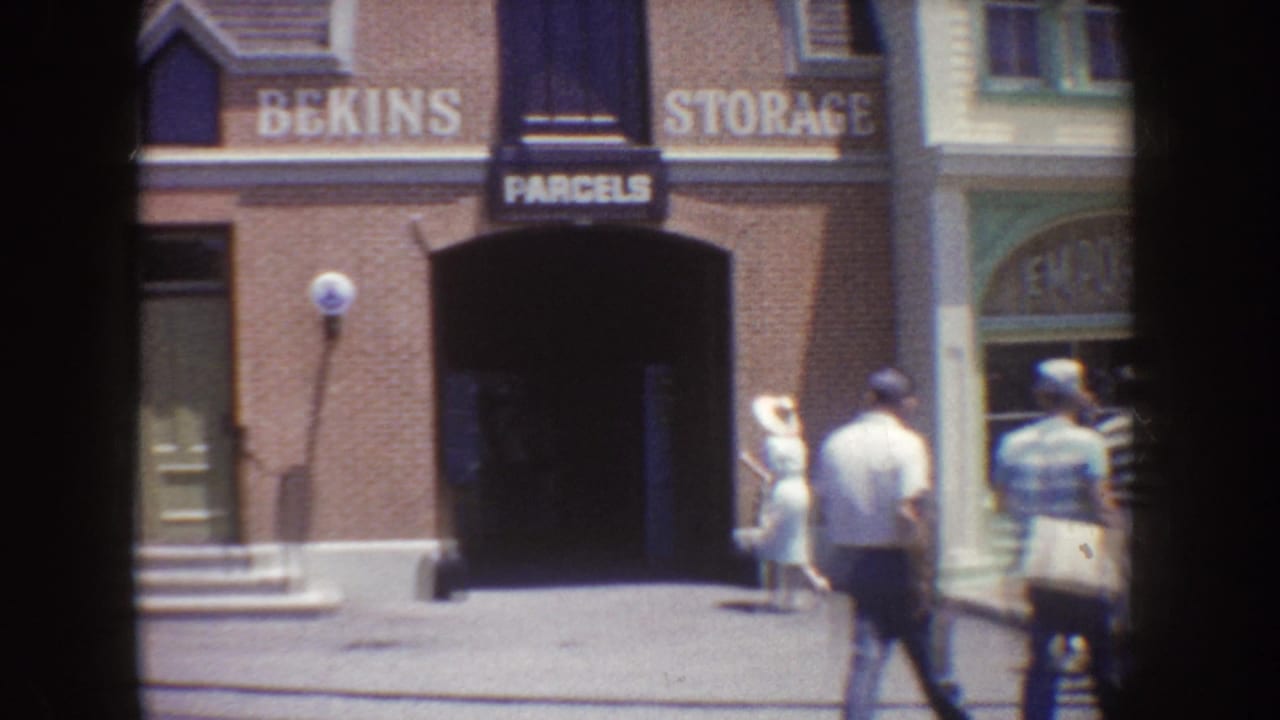 1962 Disneyland home-movie frame showing visitors walking outside a brick Bekins Storage and Parcels storefront.