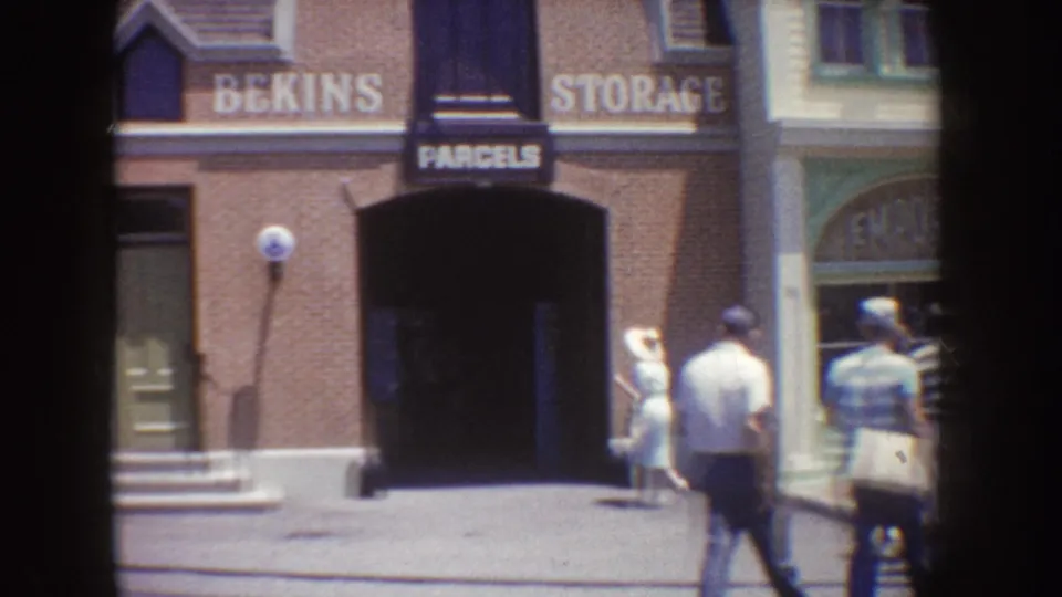 1962 Disneyland home-movie frame showing visitors walking outside a brick Bekins Storage and Parcels storefront.