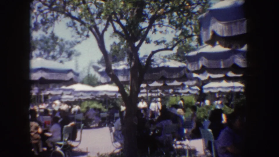 1962 Disneyland home-movie view of shaded outdoor dining with striped umbrellas, trees, and park guests at tables.