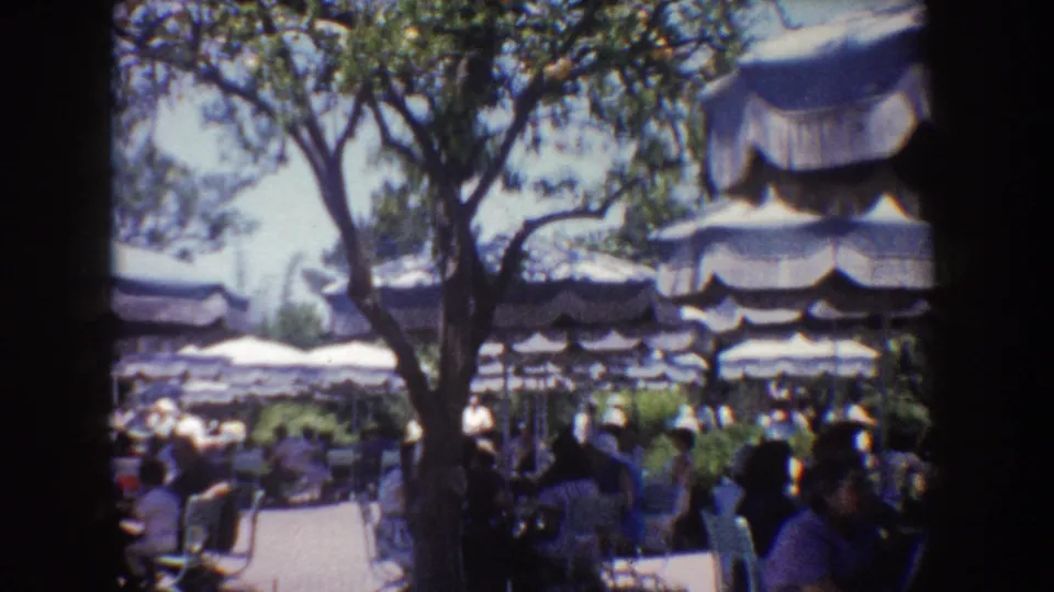 1962 Disneyland frame showing visitors gathered at tables beneath layered umbrellas and tree shade.