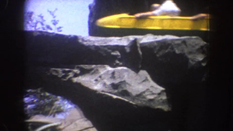 1962 Disneyland visitor footage shows a yellow roller coaster train moving past artificial rock ledges with sky visible behind it.