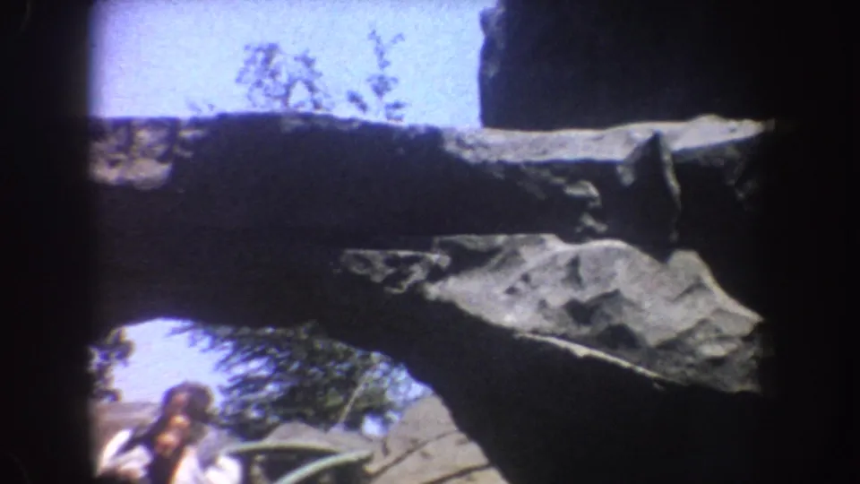 1962 Disneyland home-movie frame with artificial rockwork, sky, greenery and a partial lower-edge glimpse of the ride area.
