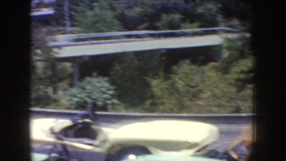 1962 Disneyland frame with a yellow ride car in the foreground, landscaped planting, railings, and an elevated bridge or track behind it.