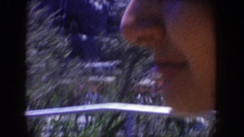 Close 8mm Disneyland frame with part of a rider at the edge of the frame, a metal rail, and blurred trees in the background.
