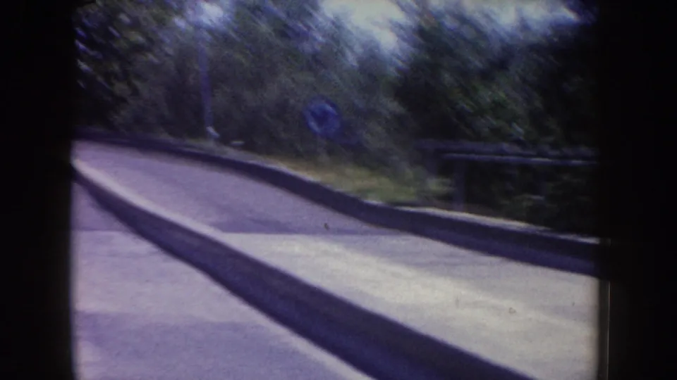 Forward-looking ride view at Disneyland in 1962, showing a curved road, trees, curb, and small blue circular sign.