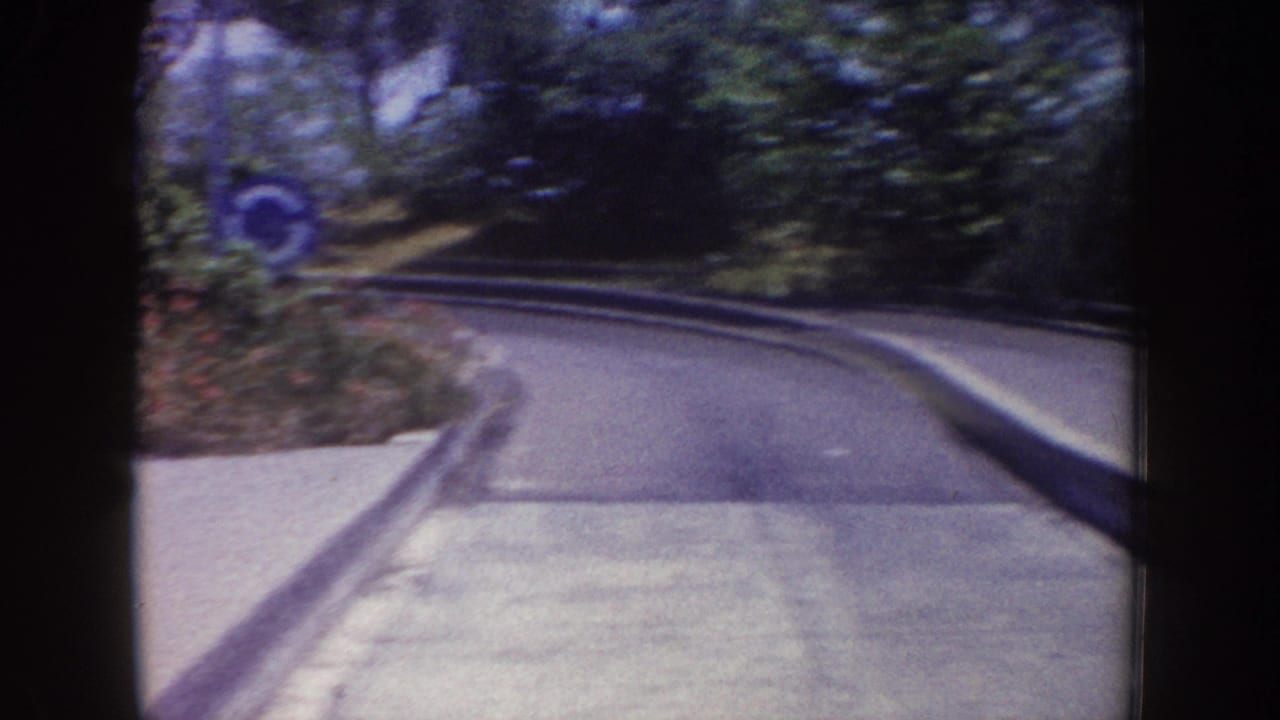 1962 Disneyland home-movie frame showing a curved paved ride track with trees, low barriers, and a blue circular sign nearby.