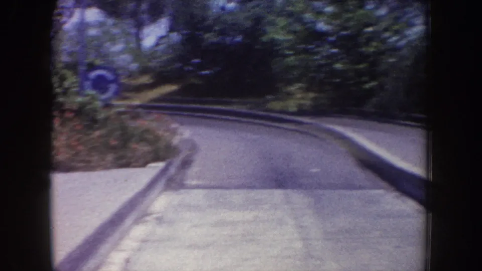 1962 Disneyland home-movie frame showing a curved paved ride track with trees, low barriers, and a blue circular sign nearby.