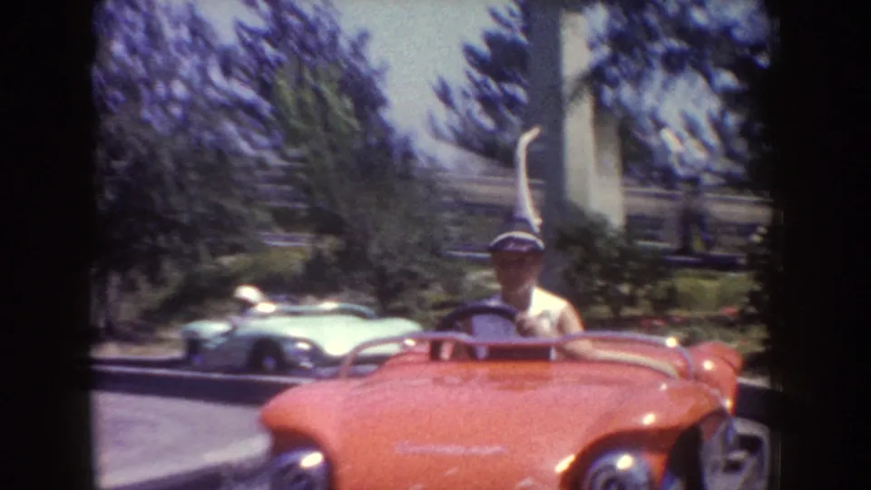 1962 Disneyland frame showing a hat-wearing guest in a red open ride car, with other small cars and trees behind.