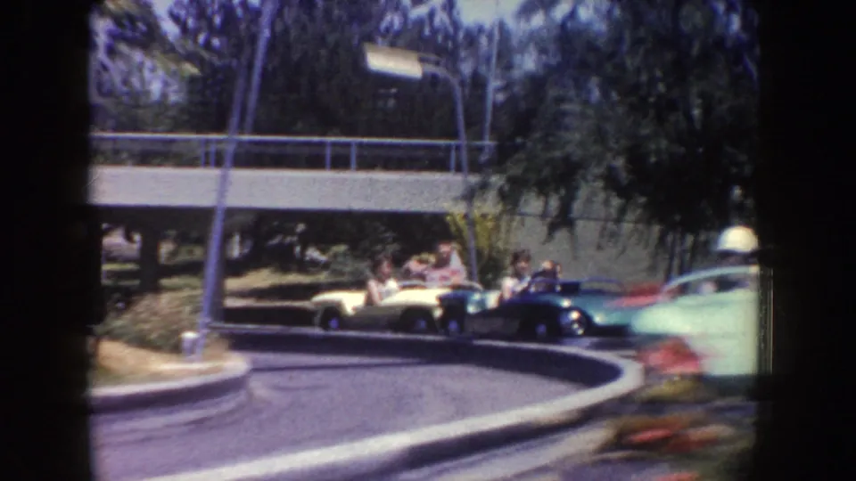 1962 Disneyland home-movie frame showing multiple guests in colorful small cars moving around a curved attraction track.