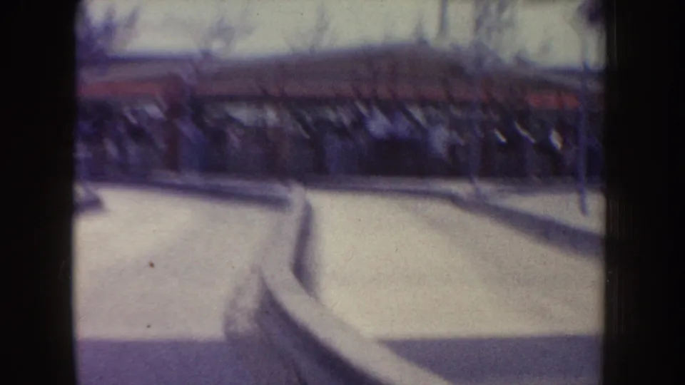 1962 Disneyland frame showing a curved paved ride track, low curb, landscaping, and a distant structure in soft 8mm focus.