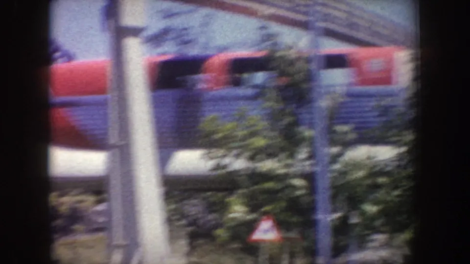 Close side view of the Disneyland monorail passing behind a window edge and greenery, with passengers faintly visible through dark windows.