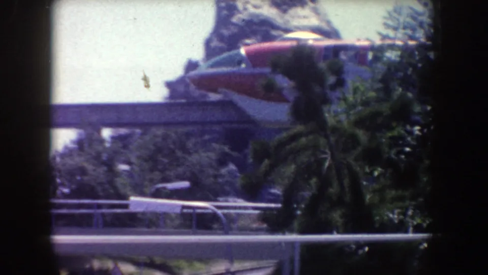 Red-and-silver Disneyland monorail travels on an elevated guideway above railings, trees, and landscaped rockwork in the 1962 footage.