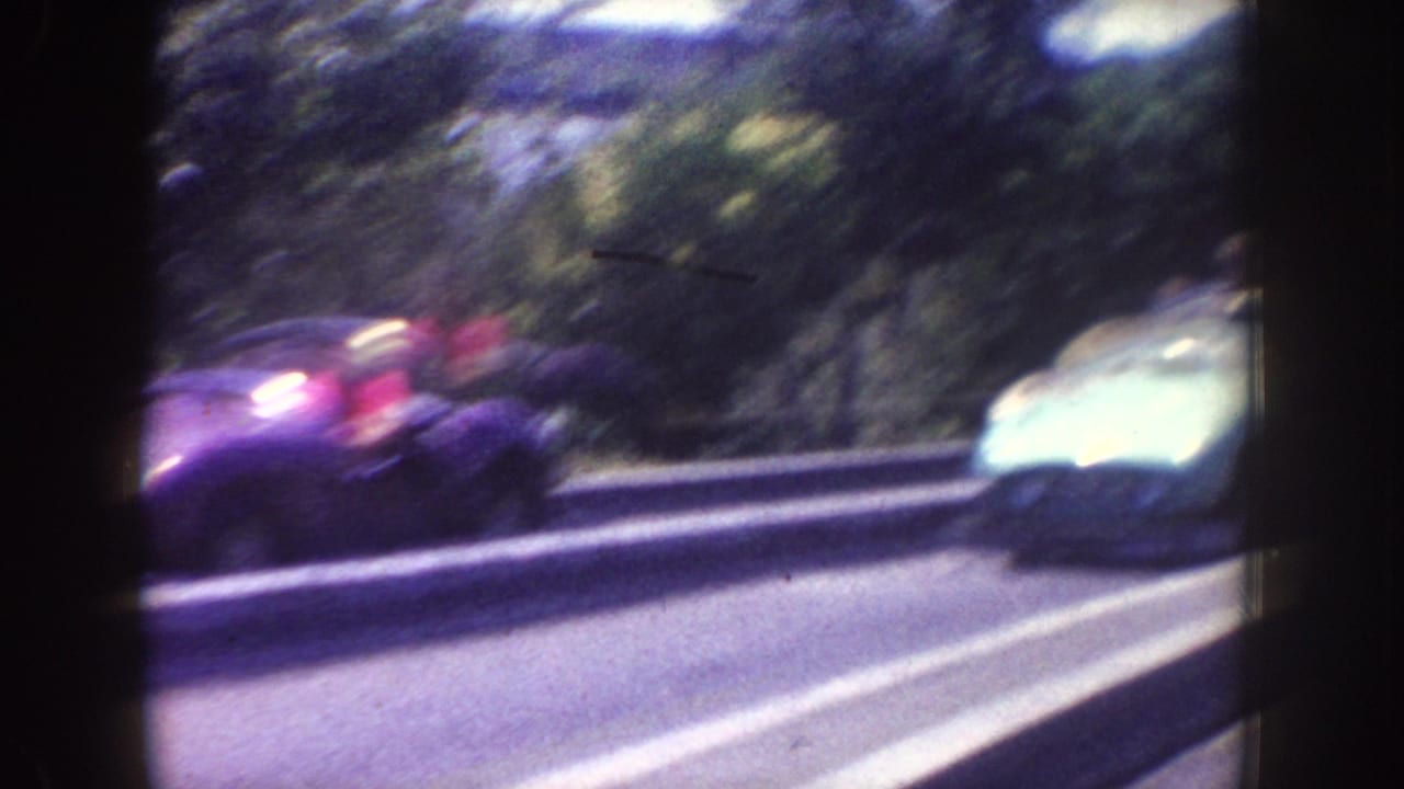 Vintage cars pass along a curved road behind a guardrail.