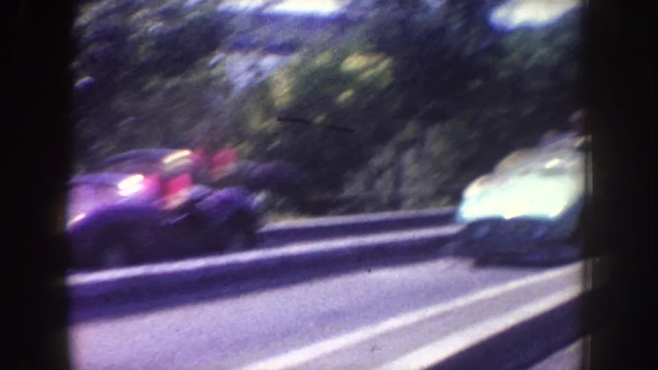 A blurred 1962 Disneyland road view shows vintage cars moving past trees and a roadside barrier from a vehicle or ride perspective.