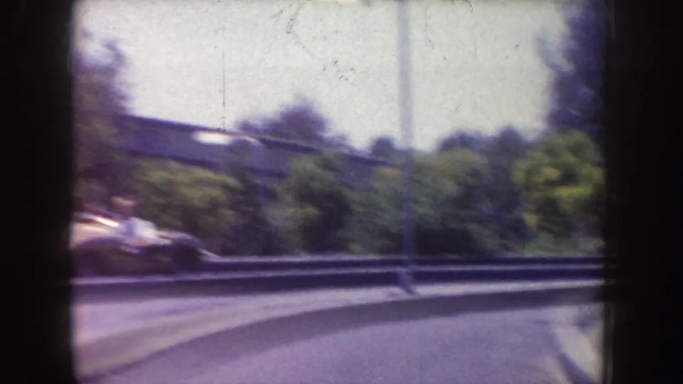 Soft 8mm frame of a curving Disneyland road or track area with trees, a guardrail, and a blurred vintage car at the edge.