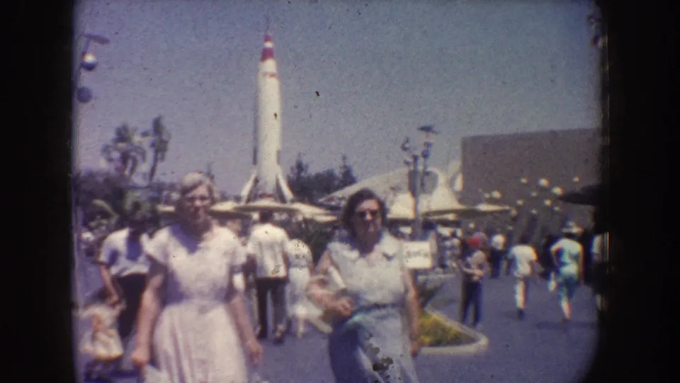 Visitors walk through Disneyland, California, near palm trees and a tall white rocket-like attraction structure in a faded 1962 home-movie frame.