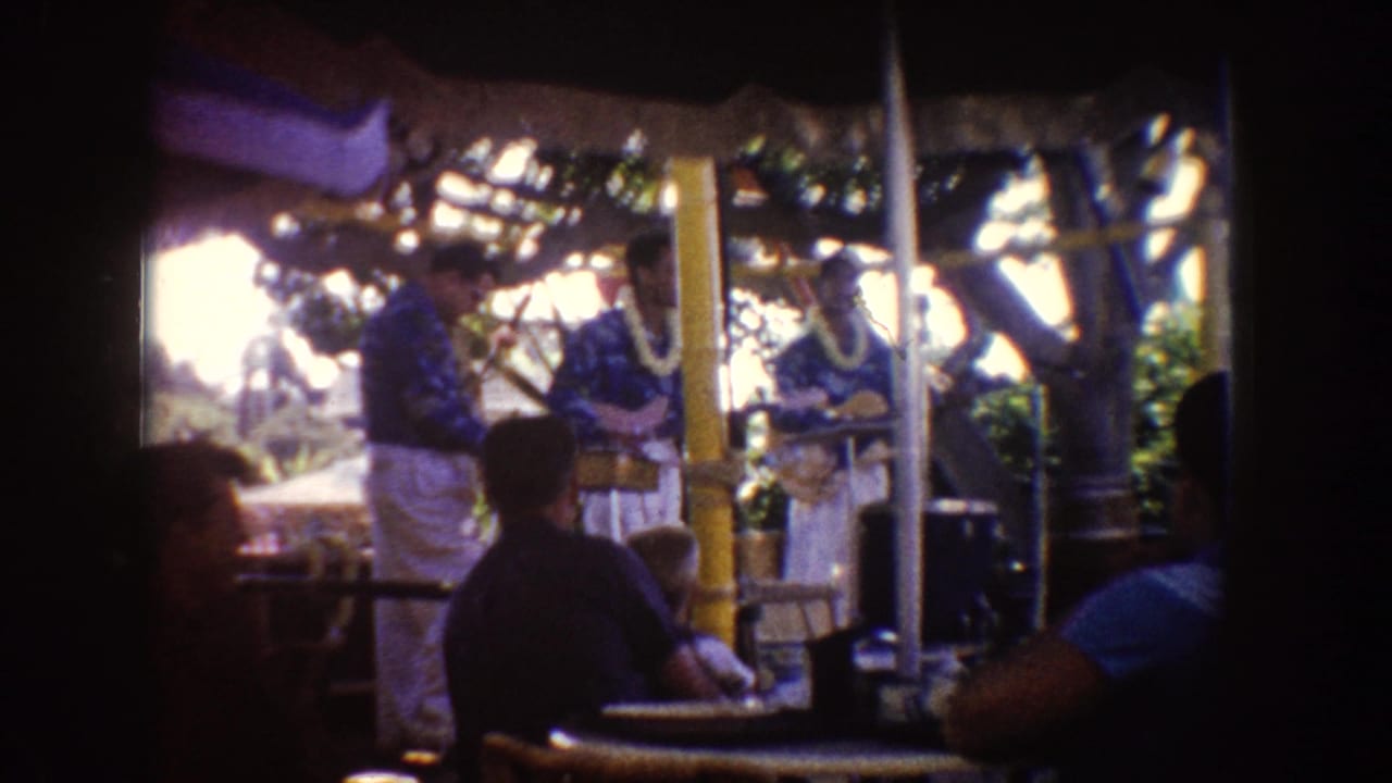 Hawaiian-style musicians perform under a thatched outdoor shelter while visitors sit nearby.