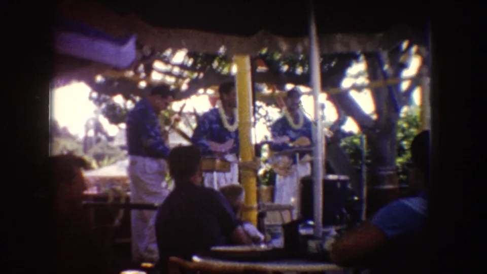 1962 Disneyland home-movie frame showing Hawaiian-style performers with leis under a thatched shelter as seated visitors watch.