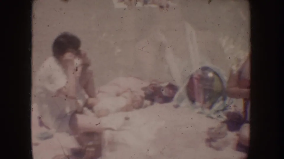Soft, overexposed 1962 home-movie frame showing a seated adult near a reclining figure or object, with film dust and faded color.