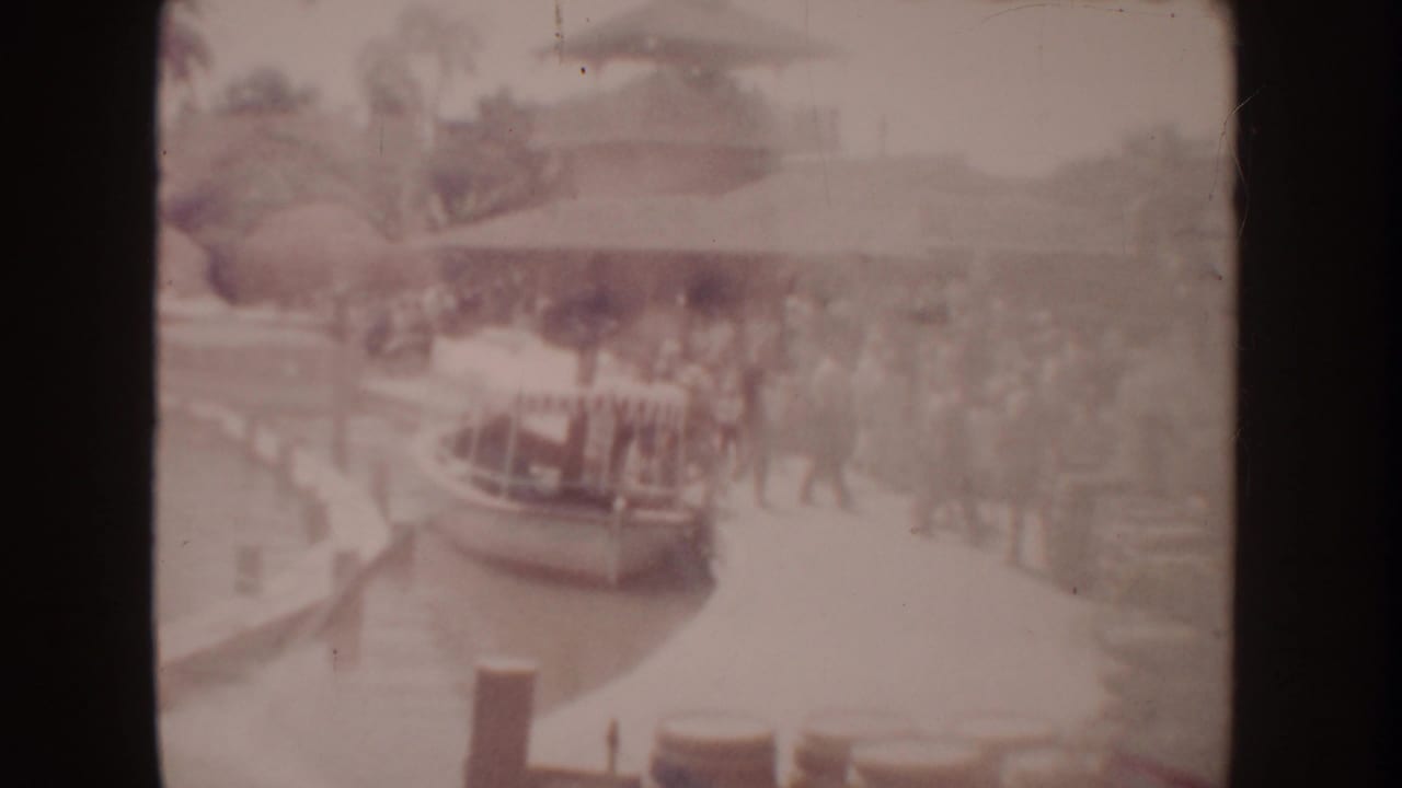 1962 Disneyland home-movie frame showing a distant crowd near a canal-side boat loading area with trees and railings.