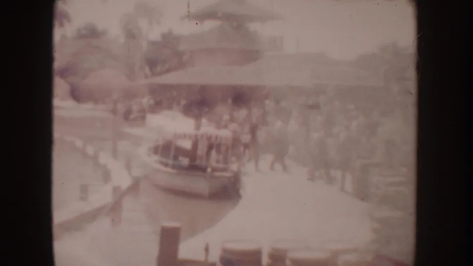 1962 Disneyland home-movie frame showing a distant crowd near a canal-side boat loading area with trees and railings.