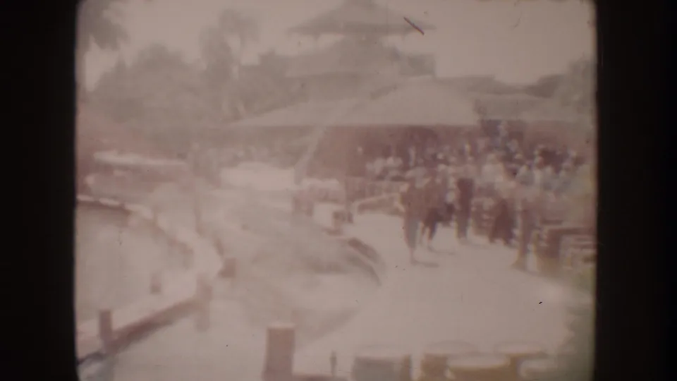 1962 Disneyland home-movie frame with visitors along a canal-side attraction area, small boats, trees, and themed building forms.