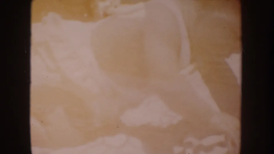 Faded 8mm frame from the 1962 Disneyland home-movie clip, showing a baby in light overalls lying on a pale blanket.