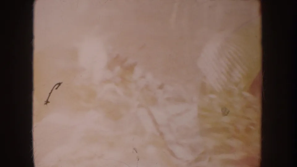 Faded 8mm frame showing a blurred blanket surface and white clothing or fabric, with film grain and scratches visible.