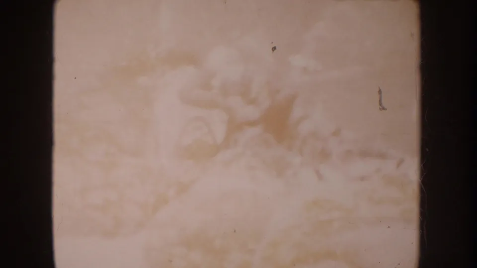 Soft 1962 home-movie frame showing a pale blanket and indistinct infant-like form, heavily faded with dust and scratches.