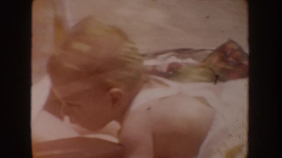 1962 Disneyland home-movie frame of a toddler in overalls crawling or leaning beside a low rocky edge.