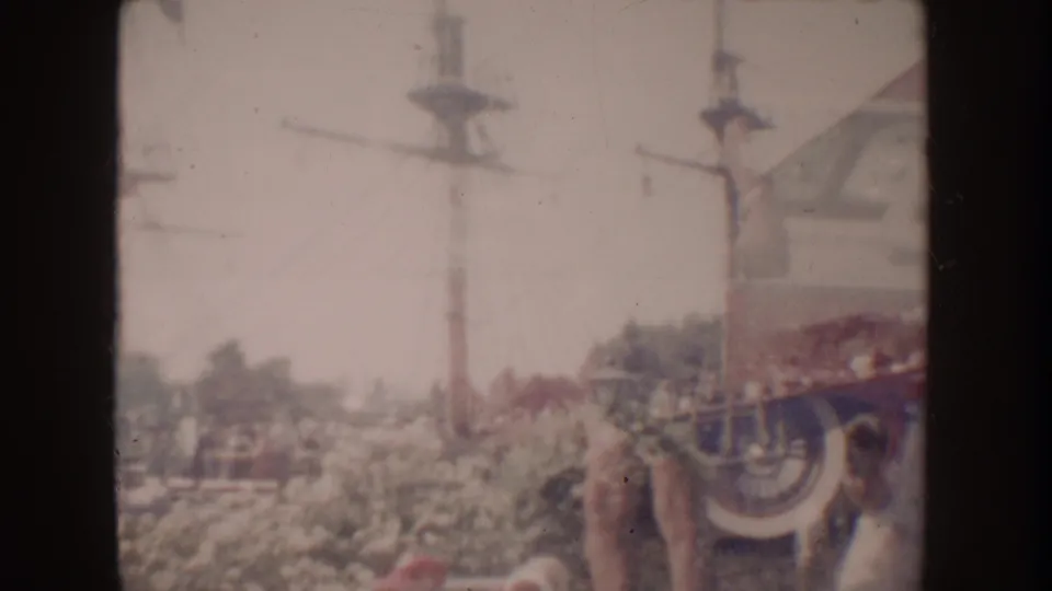 1962 Disneyland frame with transmission poles, overhead wires, landscaping, distant people, and a partial park structure at right.