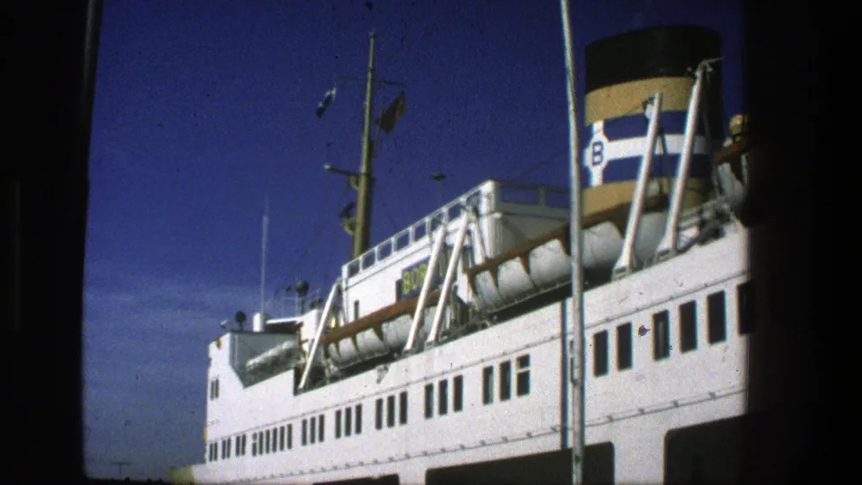 1975 Denmark ferry view with the white passenger deck, rows of dark windows, mast rigging, small flags, and blue and white striped funnel.