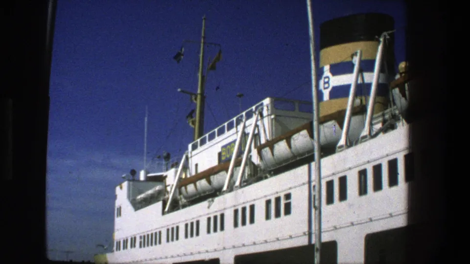 Dock-level 8mm frame of a Danish ferry, showing white decks, railings, lifeboat supports, mast, flags, and a striped funnel marked B.