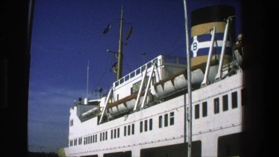 Late clip frame from 1975 Denmark showing the same white ferry at port, with windows, mast, flags, deck equipment, and striped B funnel.