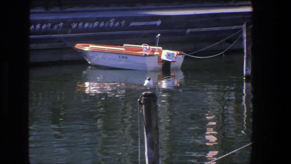 1975 Denmark harbor frame showing a small motorboat tied near a concrete quay, with a wooden post and water reflections in front.